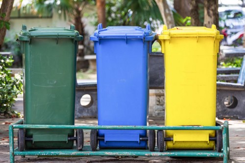 Three wheeled rubbish bins are lined up outdoors on a paved surface, each with a distinct colour and lid: the left bin is green with a closed lid, the middle bin is blue with a slightly open lid, and the right bin is yellow with a closed lid. The bins are made of durable plastic with textured surfaces and sturdy wheels for mobility. They are positioned in front of a black metal fence and some greenery, including trees and plants, suggesting an outdoor setting possibly on a driveway or communal area in Hitchin. The bins are clean and appear ready for rubbish collection, indicative of a waste management service by Commercial Waste Hitchin. The scene is lit naturally, with sunlight casting soft shadows, and the environment appears quiet and well-maintained, reflecting typical waste disposal arrangements for residential or commercial premises within the local postcode area.