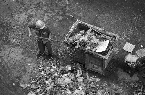 A person wearing a safety helmet and work clothes is sweeping up a pile of mixed waste and debris that has spilled onto a paved surface next to a large metal dumpster filled with various rubbish materials. The debris includes crumpled paper, plastic, and other refuse, with some waste scattered on the ground surrounding the dumpster. The scene appears to be outdoors, possibly on a driveway or service area, with a textured asphalt surface visible beneath the waste. The background shows the edge of the dumpster, which is positioned near a wall or building, and a small wooden pallet or crate is situated nearby. The lighting suggests natural daylight, emphasizing the industrial and utilitarian setting, consistent with rubbish removal or waste management activities typical in Hitchin or surrounding areas. The image reflects the sort of waste collection situation managed by Commercial Waste Hitchin, a company providing rubbish clearance services in the local area.