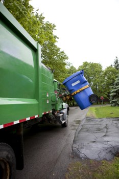 A large green commercial waste collection vehicle parked on a residential street, with its rear compartment open, exposing an array of waste bins and containers inside. The vehicle's exterior has a smooth, glossy finish with reflective red and white safety markings along the lower edge. Toward the rear of the truck, a blue wheeled bin is being loaded or unloaded, partially tilted with its lid closed, positioned on the asphalt driveway or roadway. The background shows a suburban setting with trees, grass, and a paved area, suggesting the location is within a neighbourhood. The environment appears overcast with diffused daylight, and the scene captures the process of rubbish removal, consistent with services provided by Commercial Waste Hitchin for local waste management in the Hitchin area near SG postcode region.