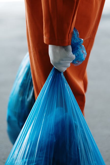 A sanitation worker in a yellow high-visibility vest is standing behind a large red waste collection vehicle parked on the side of a street. The worker is leaning forward, using a tool to unload or cl