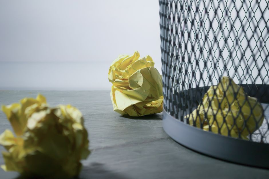 Close-up view of crumpled yellow paper balls scattered on a grey, textured surface with a metal waste bin featuring a diamond-shaped grid pattern on the right side of the image. The paper balls are ir